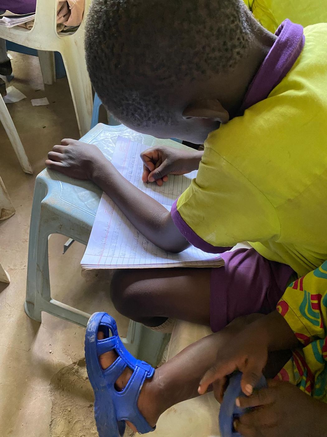 A young pupil in a lime and purple uniform bent over an exercise book resting on a plastic stool, writing carefully