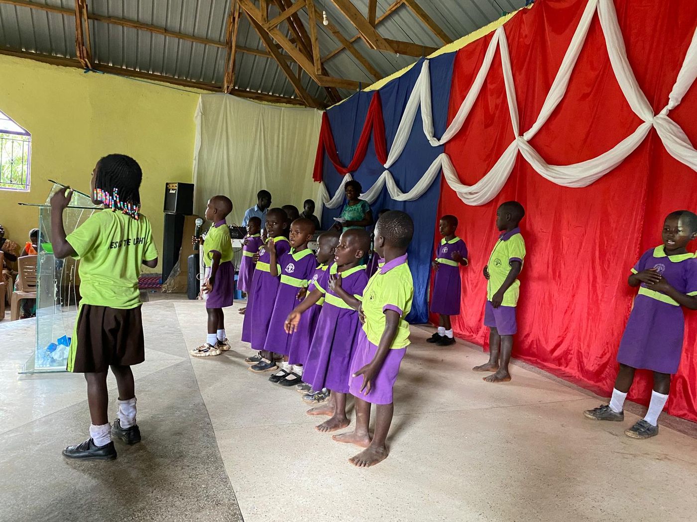 A line of young children in purple and lime school uniforms singing at the front of a church, led by an older pupil