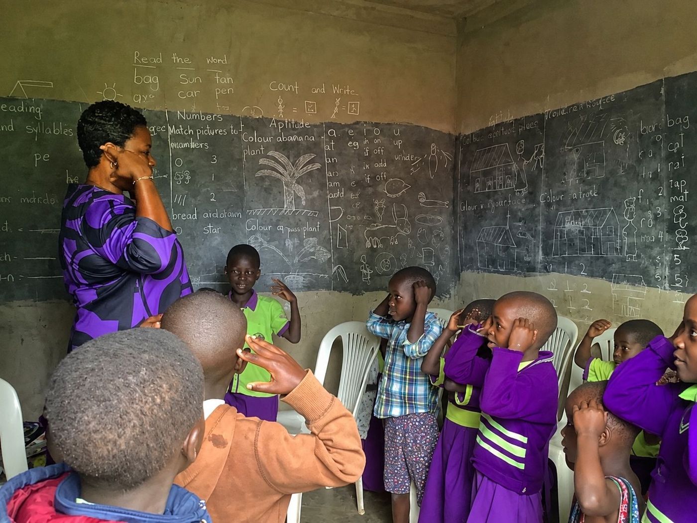 A teacher standing beside blackboard walls covered in handwritten reading and counting exercises, with young pupils in purple and lime uniforms responding