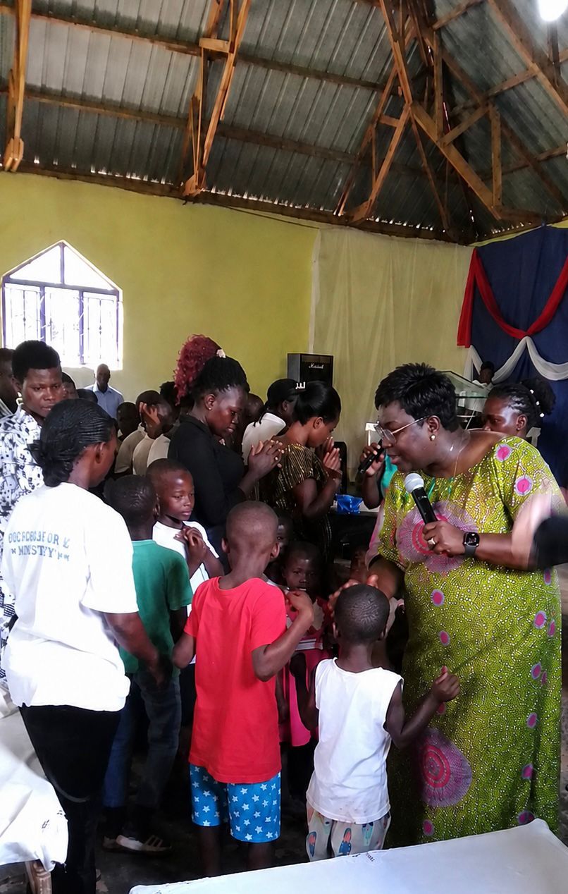 Rev. Josephine Harrison praying into a microphone while children and adults stand with hands raised and heads bowed