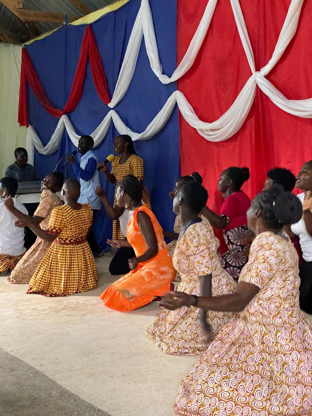 Women in patterned dresses kneeling on the floor in worship while a worship leader sings from the platform